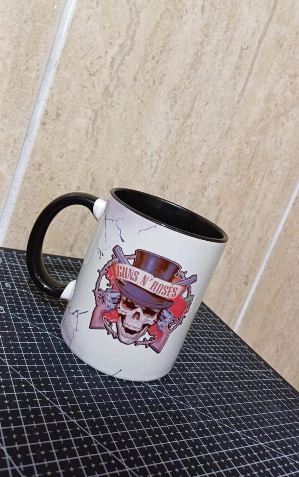 Caneca branca com interior e alça pretos, fundo marmorizado e estampa de caveira com cartola, rosas e pistolas da banda Guns N' Roses