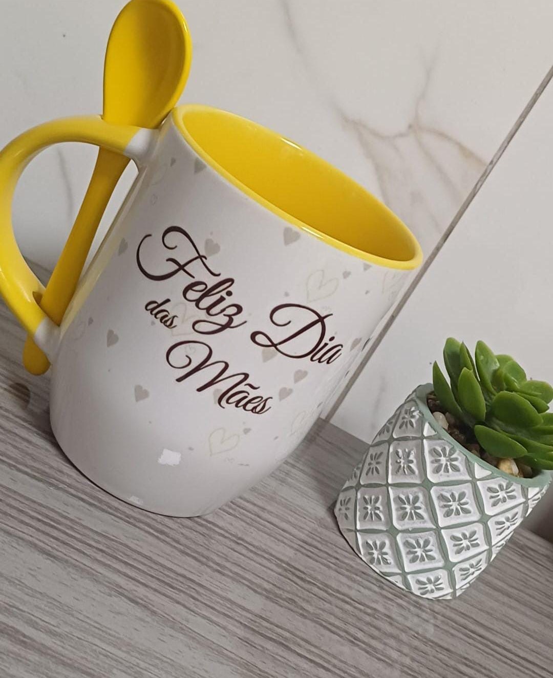 Caneca branca com interior, alça e colher amarelos, corações e a frase Feliz Dia das Mães