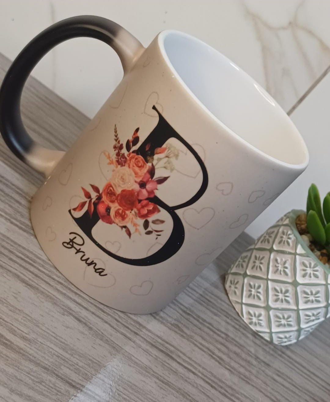 Caneca fosca em tom areia com alça preta, letra B grande com rosas em tons terrosos, corações e o nome Bruna