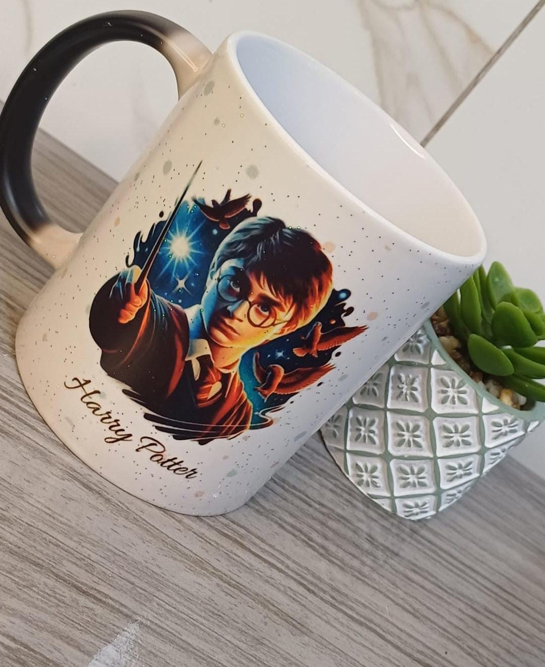 Caneca clara com respingos, alça preta e estampa de um jovem bruxo de óculos com varinha, corujas e o texto Harry Potter