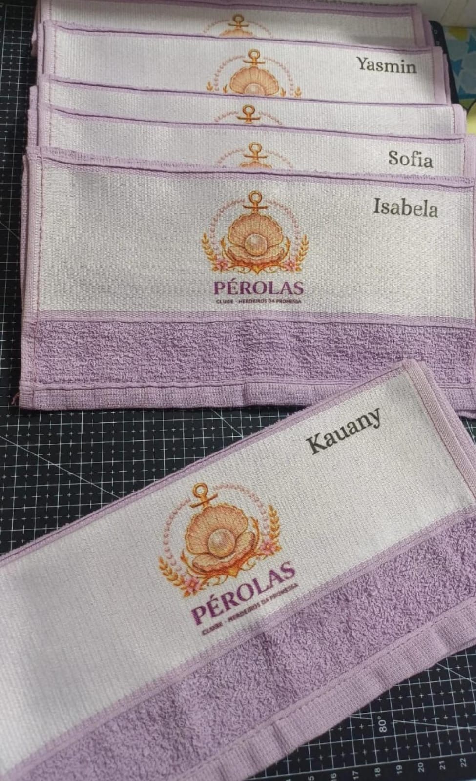 Toalhinhas lilás com a logo do clube Pérolas, uma concha com pérola e âncora, e os nomes Yasmin, Sofia, Isabela e Kauany